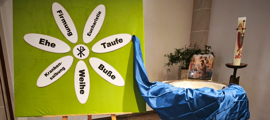 Taufbrunnen mit Schautafel, im Kreis angeordnet die Aufzählung: Taufe, Buße, Weihe, Krankensalbung, Ehe, Firmung, Eucharistie